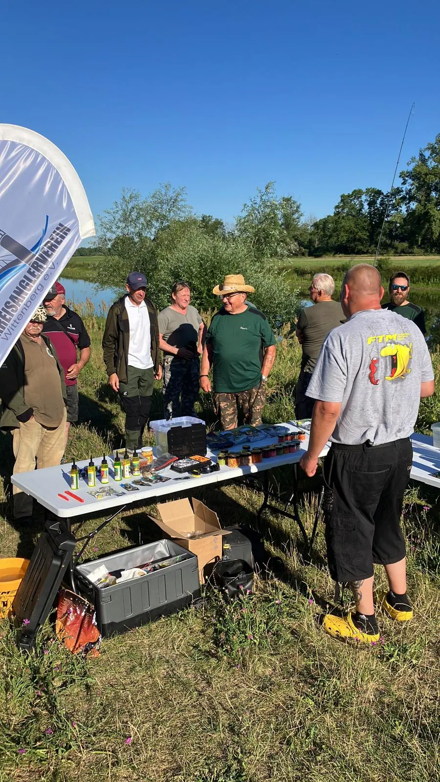 Gerätetisch mit Futterzusätzen, Aromen und Zubehör von Fishing Tackle Max, umringt von Seminarteilnehmern am Wasser