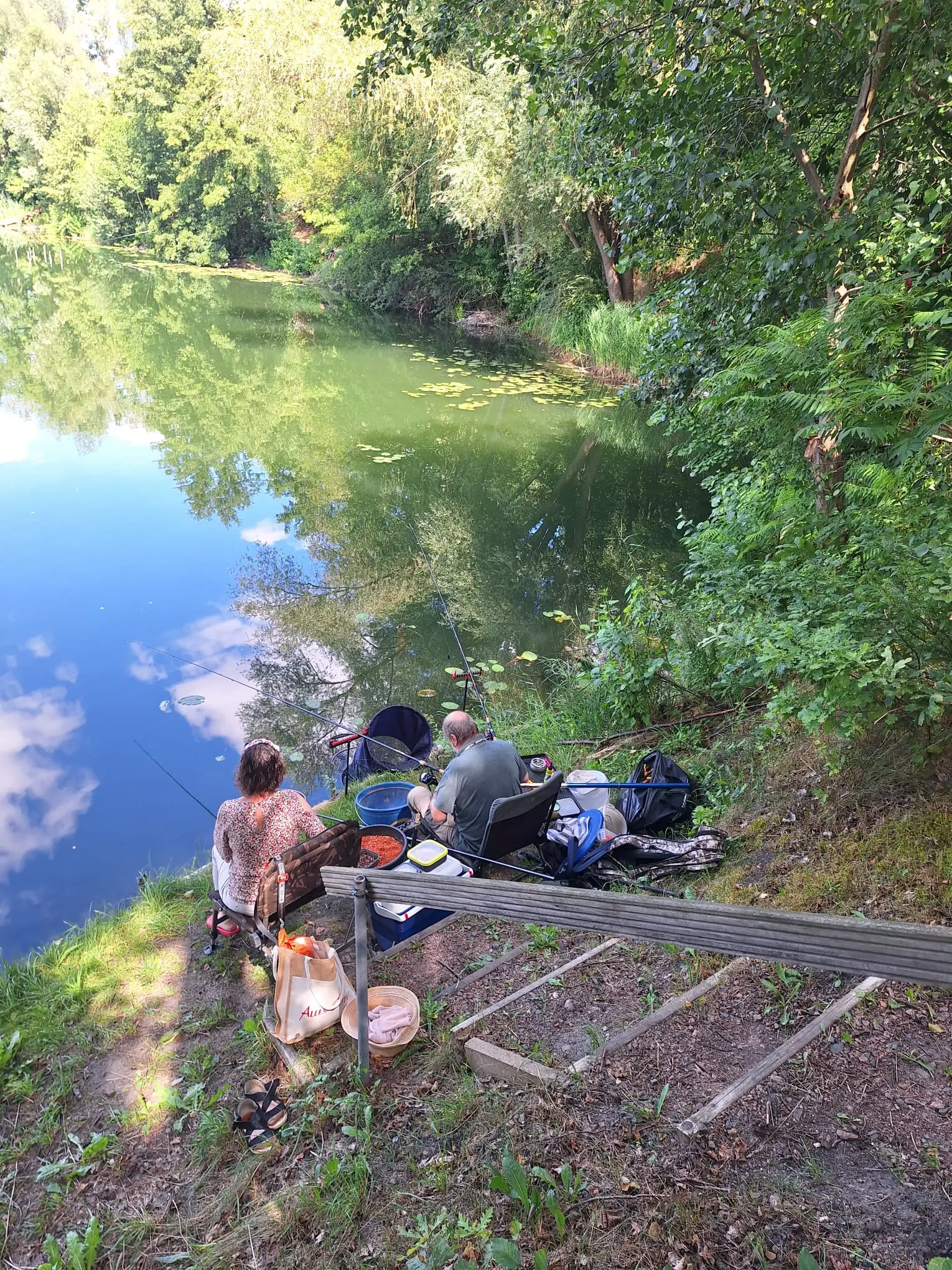 Ein Paar sitzt mit Ruten und Ausrüstung am Ufer des Gewässers, im Wasser spiegeln sich Himmel und Bäume
